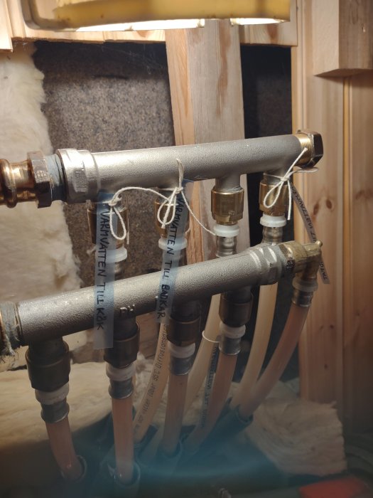 Vattenrör och fördelare med etiketter och en pluggad mässingsdel i ett badrum under renovering.
