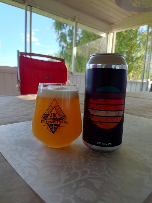 Ölglas med skumkrona bredvid en burk Alefarm Double IPA på ett bord på en veranda.