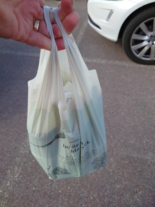 Hand håller en vit plastpåse med text framför en parkerad bil.