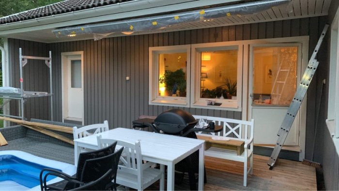 Hus med ny målning delvis färdig, stege mot vägg, terrass med grill och möbler.