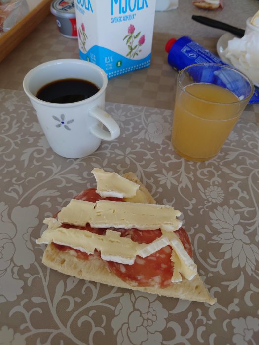 Frukost med kaffe, apelsinjuice, och en macka med tomat och ost på ett mönstrat bord.