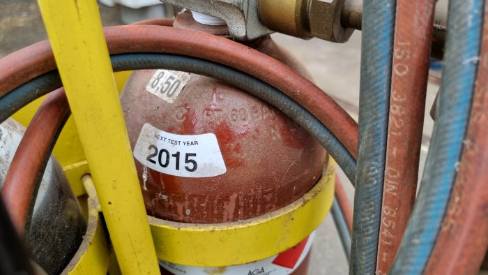 Närbild på en gammal röd gasflaska med slangenätverk och en etikett för nästa testår 2015.