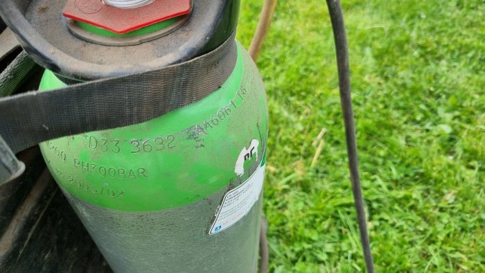 Grön gasflaska utan skyddskåpa med synliga etiketter och märkningar, står i ett gräsområde.