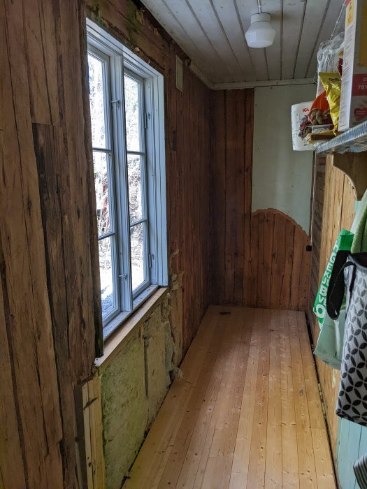 Ett hörn av ett gammalt torp med stående träplank på väggarna, ett fönster och saknad plankbit under fönstret.