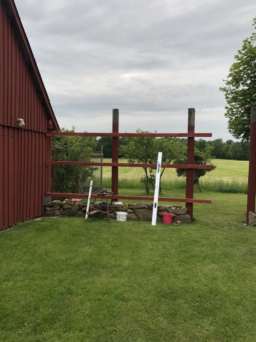 Delvis byggt staket med horisontella plankor, steinhägn framför, byggredskap på gräsmatta.