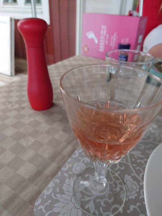 Ett glas rosévin på ett bord med blommigt mönster, en röd kvarn och en rosa låda i bakgrunden.