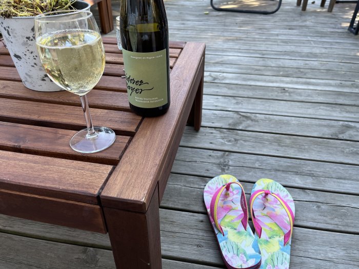 Ett glas vin och en flaska vid träbänk på däck, bredvid färgglada flip-flops.