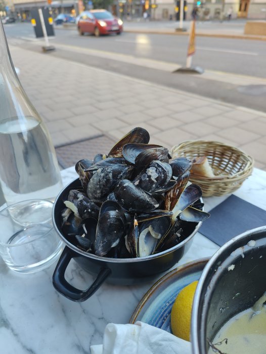 En gryta med moules marinières och en korg med bröd på ett bord vid en gata.