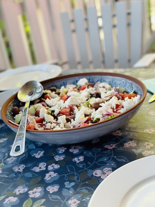 Grekisk sallad i en blå skål på bord med blommigt duk, utomhusmiljö.