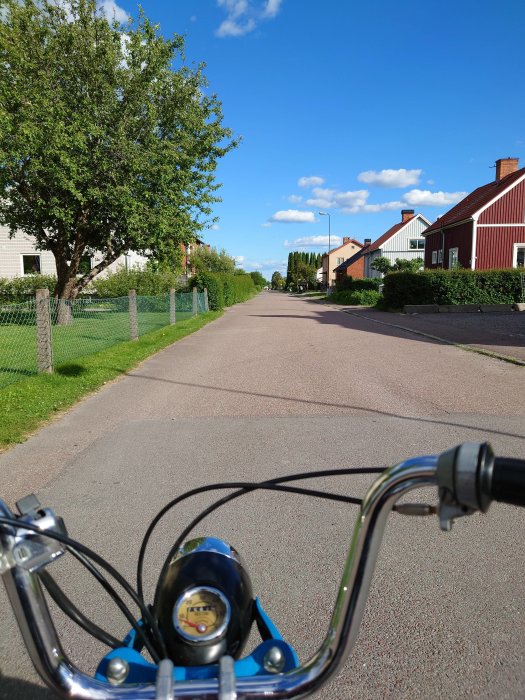 Vy från en cykel som rör sig längs en lugn bostadsgata med träd och enfamiljshus under en klar dag.