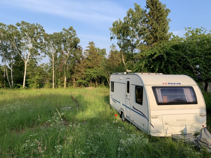 Husvagn parkerad på grön äng med träd i bakgrunden under klar himmel.