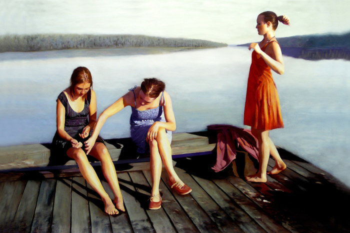 Tre unga kvinnor vid en sjö; två sitter på en brygga medan en står och tittar ut över vattnet.