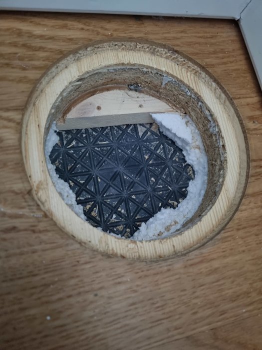 Urtaget i trägolv visar isolering och svart plast, troligen ångspärr, i en fuktkontrollöppning.
