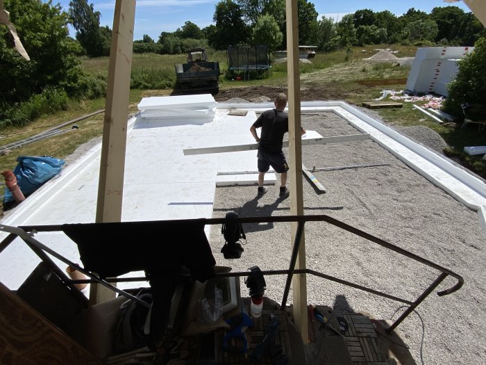 Person på en grund med utlagd cellplast och armeringsnät, arbetet pågår, byggmaterial och grävmaskin i bakgrunden.