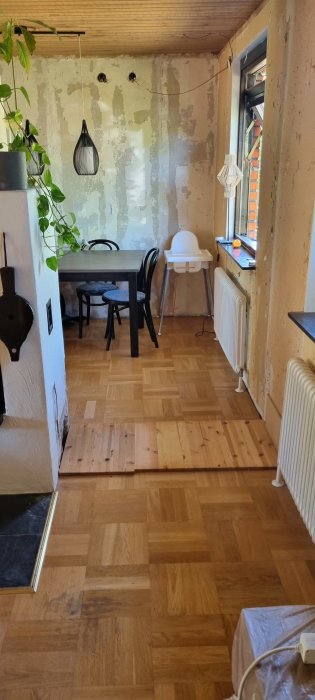 Rum under renovering med blandat parkett- och plankgolv, avskalade väggar och en matplats med stolar och bord.