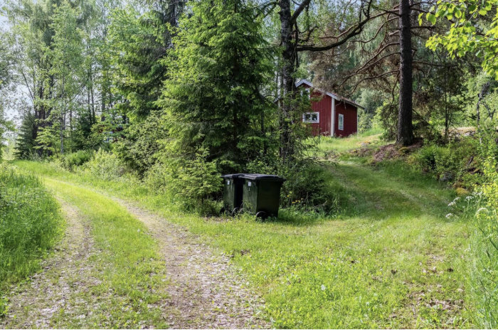 Smal grusväg leder till en uppfart med en röd stuga och gröna soptunnor i naturlig omgivning.