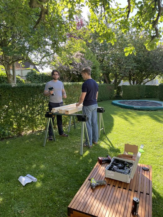Två personer arbetar på ett träbyggprojekt i en trädgård, med verktyg och material synliga.