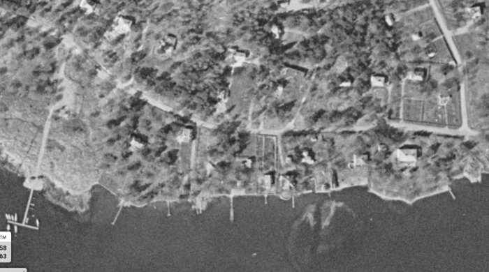 Svartvitt flygfoto från 1960-tal på kustlinje med privata bryggor och egendomar.