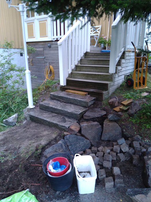 Nybyggd stentrappa vid en trädgård med trappsteg av grova stenblock, verktyg och hinkar synliga.