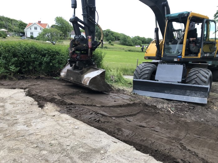 Grävmaskin som jämnar ut jord och förbereder underlaget för altanbygge, med sandbädd nära bergssidan.