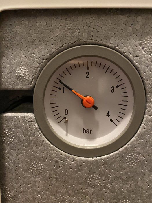 Tryckmätare med orange visare visar drygt 1 bar, monterad på en grå yta med struktur.