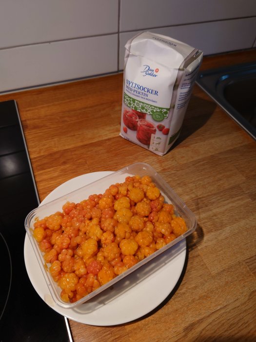 En förpackning med syltsocker bredvid en genomskinlig behållare fylld med orangefärgade bär på ett köksbord.