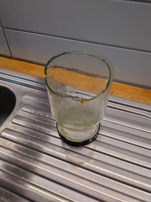 Ett använt genomskinligt dricksglas med rester och droppar av vätska på en rostfri diskbänk.