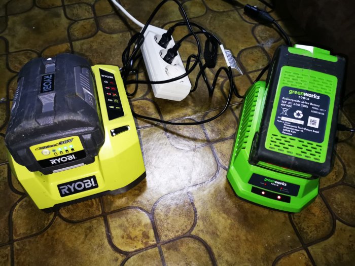 Ryobi och Greenworks batterier med laddare kopplade till ett uttag på ett brunmönstrat golv.