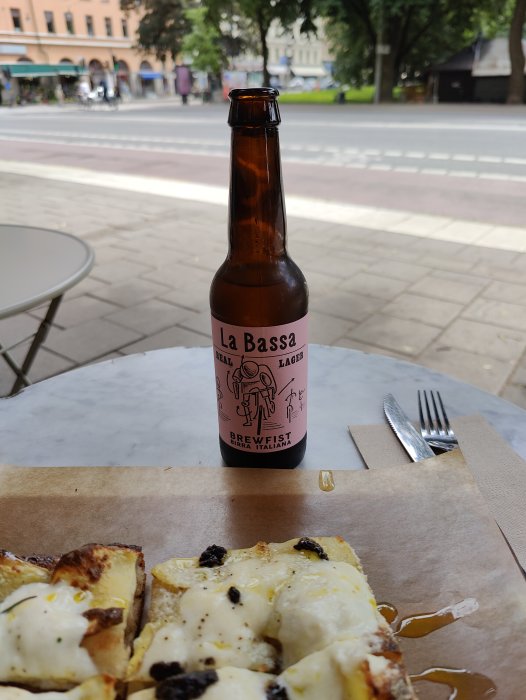 Öppen ölflaska av La Bassa Lager på bordet bredvid pizza på en uteservering.