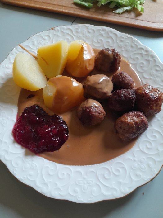 Tallrik med köttbullar, kokt potatis, brunsås och lingonsylt på ett vitt spetsmönstrat fat.