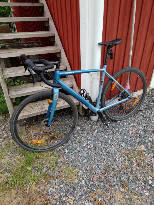 En blå landsvägscykel lutad mot en röd trävägg nära en trätrappa med grusunderlag.