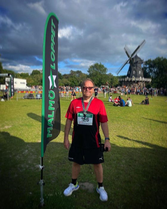 Person i löpartenue står framför Malmö Loppet-skylt med medalj, vindkvarn i bakgrunden.