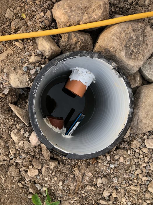 Närbild på en nyligen installerad dräneringsbrunn med anslutna rör omgiven av sten och grus.