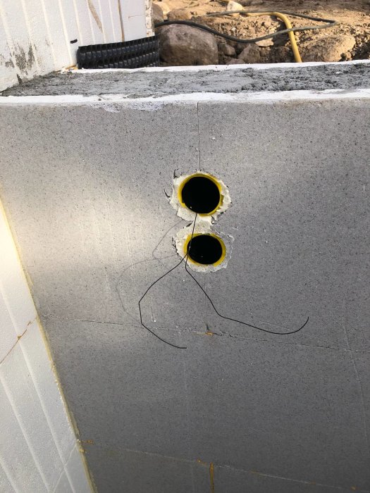 Grå betongvägg med två hål tätade med gula rörändar, omgivet av byggmaterial och grusmark.