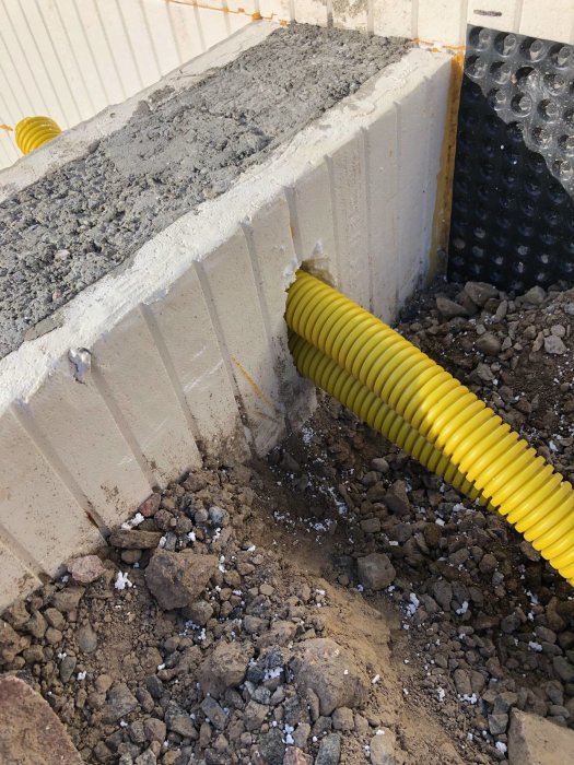 Nygjuten betongfundamentkant med gul avvattningsslang och svart dräneringsplatta vid en byggarbetsplats.