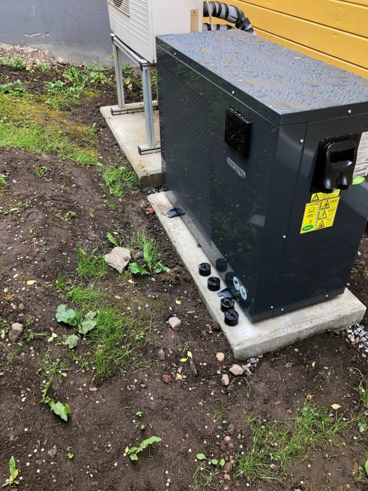 Nyinstallerad värmepump på betongfundament med anslutna rör vid husvägg och grusad mark runtom.