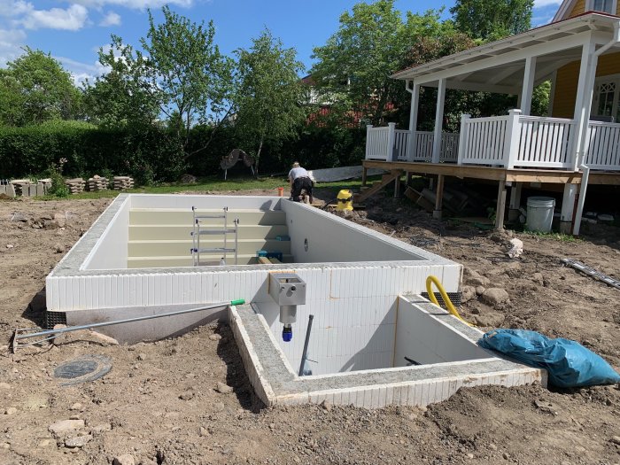 Byggarbetsplats med person som arbetar nära en ofärdig pool, med sladdar och slangar på marken.