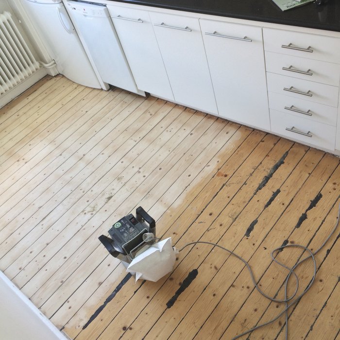 Slipmaskin på ett nött trägolv i kök med vita skåpluckor.