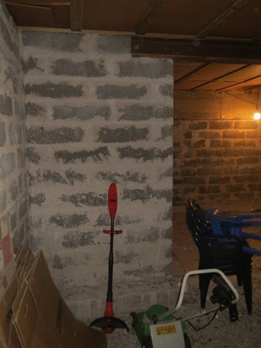 Seinäkappale autotallissa, jossa harmaa kiviseinä, punainen puutarhatyökalu ja vihreä ruohonleikkuri. Seinää on tarkoitus lyhentää.