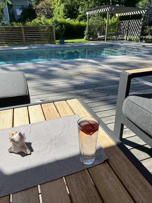 Soligt trädäck med ett glas dryck, en snäcka, och en inbjudande pool i bakgrunden omgiven av en välskött gräsmatta.