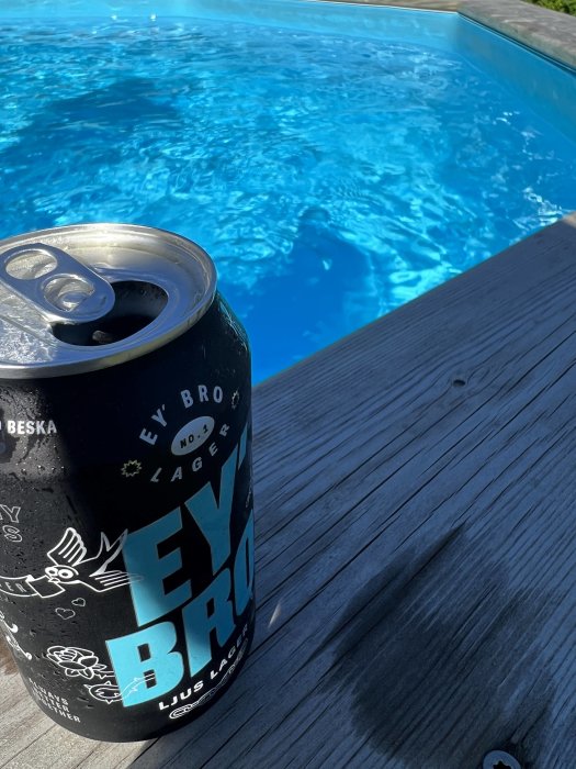 Burk med dryck på trätrall framför blå swimmingpool i solljus.