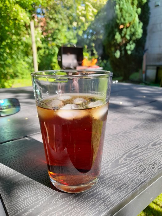 Glas med rom och cola på trädgårdsbord, grill i bakgrunden, sommardag.