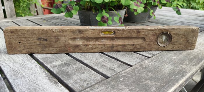 Ett slitet trävattenpass placerat på ett grått träbord utomhus, med gröna växter i bakgrunden.
