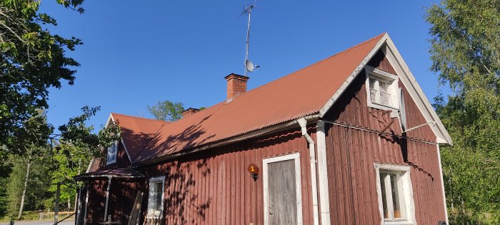 L-format rött trähus med lutande tak och skorsten, omgivet av grönska under klarblå himmel.