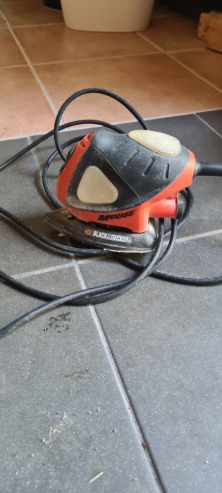 Sliten Black & Decker Slipmus elektriskt verktyg på stengolv med synlig sladd och några sprickor på dess yta.