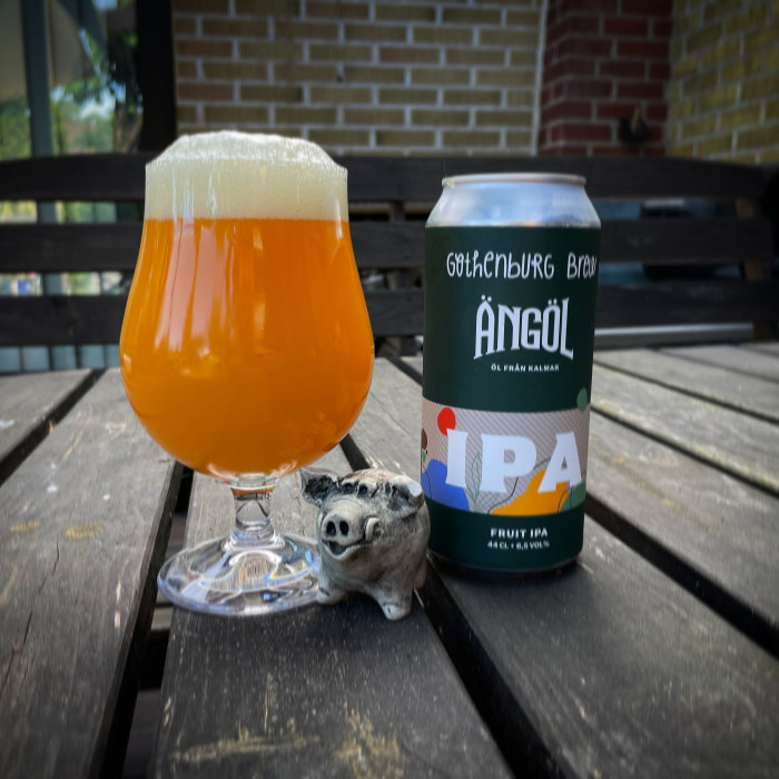 Ett glas orangefärgad öl och en burk IPA på ett träbord utomhus med en liten figur av en gris.