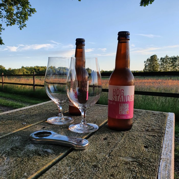 Två tomma ölglas, en öppnare och två ölflaskor på ett träbord utomhus vid skymning.