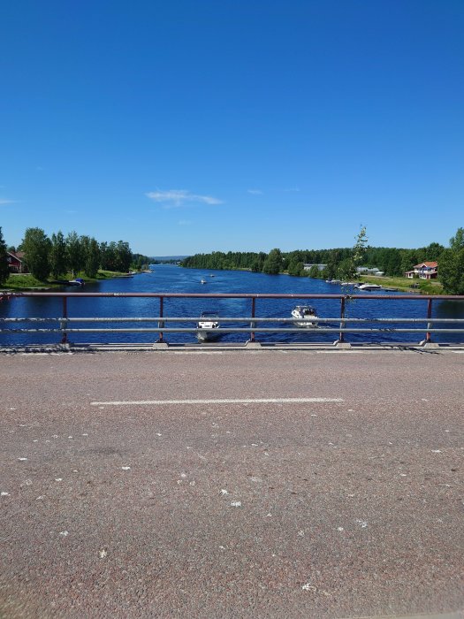 Vy över en flod från en bro i Torsång med båtar förtöjda längs stranden i soligt väder.