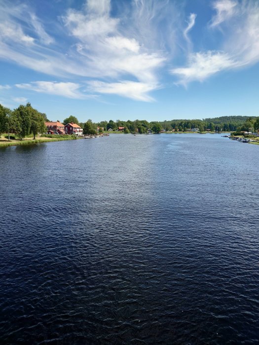 Vy över en lugn flod i Torsång med båtar och traditionella röda hus längs stranden under en blå himmel.