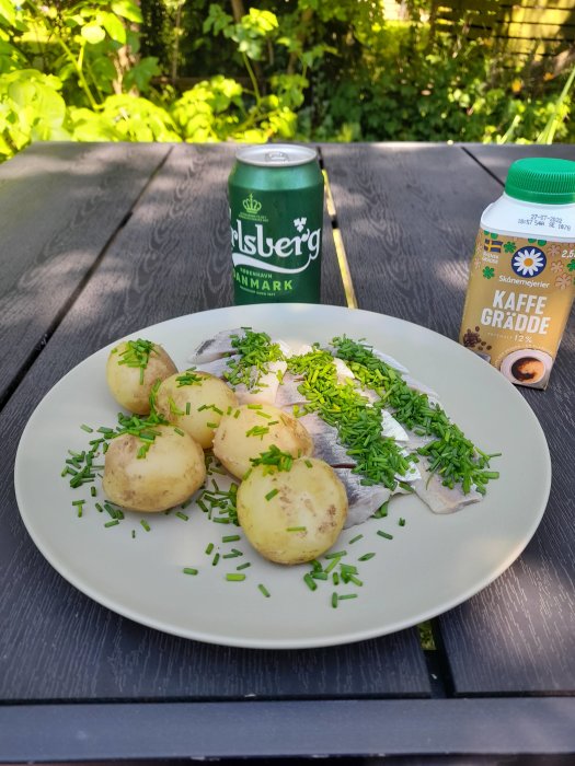 Tallrik med sill, kokt nypotatis, gräslök och en burk kaffegrädde samt ölburk på ett trädgårdsbord.